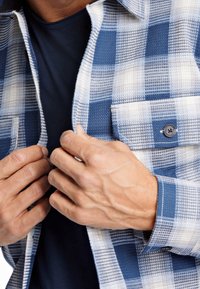 Sinivalkoruudullinen flanellipaita, jossa on nappitasku, teksturoitu kangas ja sen alla osittain näkyvä tumma aluspaidan osa.