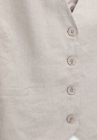 Lnianowa kamizelka w jasnym beżu z pięcioma okrągłymi jasnymi guzikami. Posiada jedną kieszeń po lewej stronie, teksturowana powierzchnia materiału.