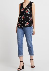 Frau in schulterfreiem schwarzen Blumenoberteil, mit umgeschlagenen blauen Jeans und schwarzen High-Heels, steht vor einem einfarbigen Hintergrund.