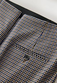 Herreblazer i et brunt, blåt og sort hounds-tooth mønster. Har en sort knap og to øverste lommer med sort foring. Tekstureret stof.