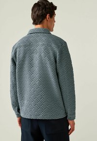Quilted-Jacke aus strukturiertem hellblauen Stoff, mit einem Zickzackmuster, Kragen und geknöpften Manschetten. Von hinten gezeigt.