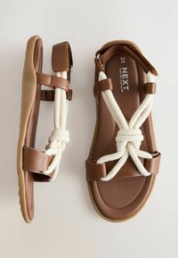 Sandales plates marron avec brides croisées en corde blanche, fermeture velcro réglable à la cheville, semelle intérieure rembourrée et marquages de taille à l'intérieur.