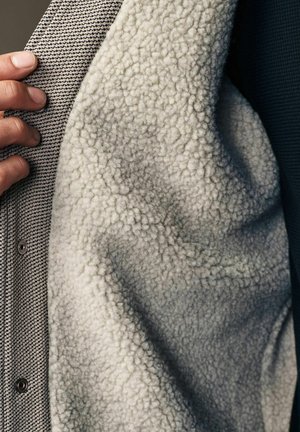 Mano che tiene aperta una giacca di lana testurizzata che mostra una morbida fodera in pile color crema, a contrasto con il tessuto esterno a motivo.