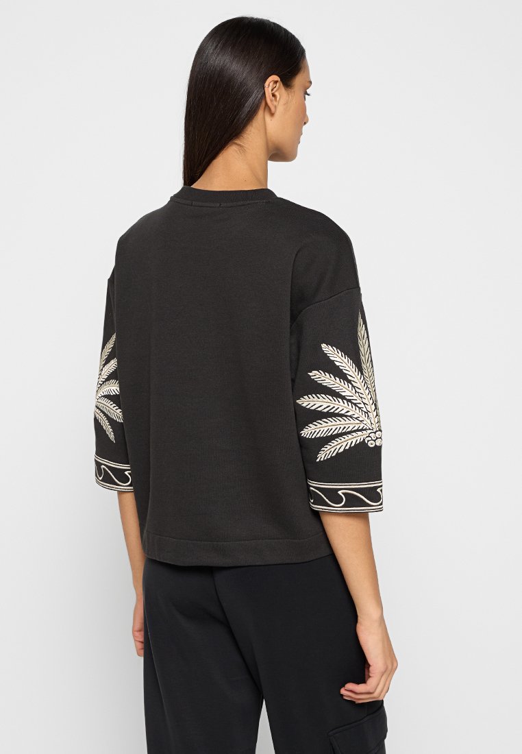 Sudadera negra con mangas tres cuartos, que presenta bordado de hojas beige en las mangas y puños decorativos. Ajuste casual y textura suave.