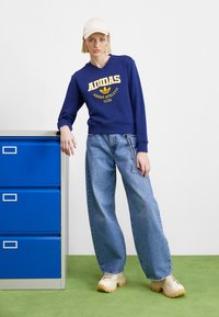 Jūrinis džemperis su geltonu Adidas logotipu, derinamas su šviesiai mėlynomis plačiomis kelnėmis ir kremiškais sportbačiais. Fone matoma mėlyna stalčių spinta.