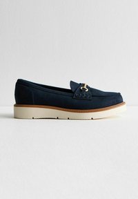 Mocassin en suede bleu marine avec un bout arrondi, des accents en métal doré et une semelle en caoutchouc couleur crème, featuring une bordure rebrodée le long du bord.