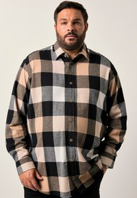 Uomo con barba che indossa una camicia a quadri oversize beige, nera e grigia, in piedi contro uno sfondo chiaro e semplice con le mani in tasca.