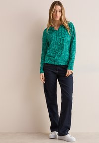 Grünes Langarmhemd mit geometrischen Mustern, kombiniert mit dunkelblauen Jeans und weißen Sneakers. Das Hemd hat einen kragenförmigen Halsausschnitt und einen lockeren Schnitt.