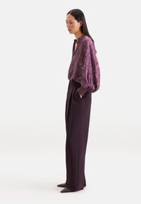 Femme portant une blouse violet à motif cachemire avec des manches ballon et un pantalon large taille haute foncé, debout les mains dans les poches devant un fond uni.