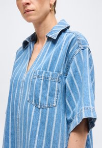 Γυναίκα που φορά μπλε και λευκό κάθετα ριγέ τζην πουκάμισο με κοντά μανίκια, τσέπη στο στήθος και γιακά ανοιχτό, που δείχνει χρυσά σκουλαρίκια κρίκους.