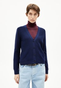 Donkerblauwe cardigan gemaakt van breiware, met een V-hals, vijf knopen aan de voorkant en geribde manchetten; gedragen over een bordeauxrode coltrui.