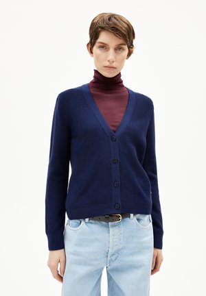 Donkerblauwe cardigan gemaakt van breiware, met een V-hals, vijf knopen aan de voorkant en geribde manchetten; gedragen over een bordeauxrode coltrui.