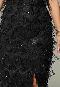 Svart klänning med en texturerad yta prydd med paljetter och pärldekorationer. Den har ett diamanthmönster och en hög slits på ena sidan.