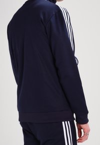 Chaqueta de chándal navy hecha de tela suave, con rayas verticales blancas en las mangas, puños acanalados y un diseño de cuello alto.