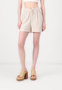 Shorts en coton beige, doux, avec taille élastique et cordon de serrage. Accompagnés de sandales à plateforme beige clair avec talons en bois et bouts ouverts.
