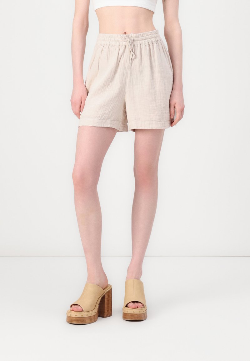 Shorts en coton beige, doux, avec taille élastique et cordon de serrage. Accompagnés de sandales à plateforme beige clair avec talons en bois et bouts ouverts.