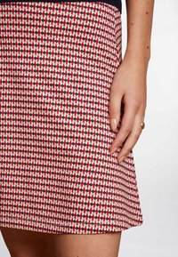 Zbliżenie dłoni z złotym pierścionkiem spoczywającej na spódnicy w geometryczny wzór w czerwonym, białym i granatowym kolorze.