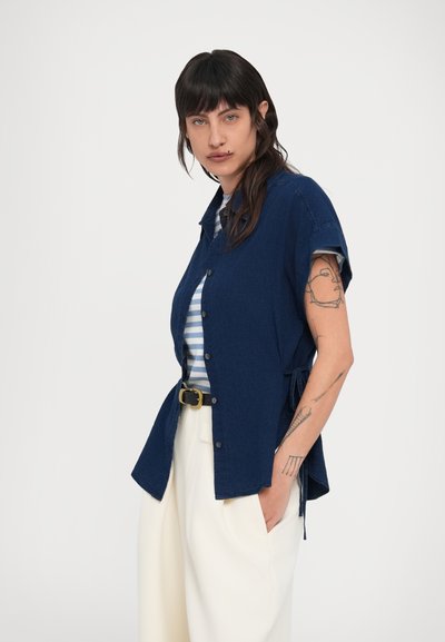 Σκούρο μπλε κοντομάνικο πουκάμισο με γιακά και κουμπιά, συνδυασμένο με ριγέ πουκάμισο από κάτω· φορεμένο με λευκό παντελόνι φαρδιάς γραμμής.