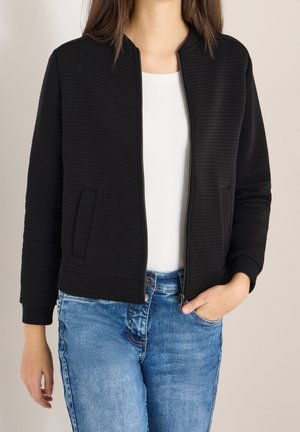 Femme portant une veste noire texturée à fermeture éclair sur une chemise blanche et un jean bleu, une main dans la poche, debout devant un fond uni.