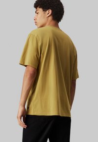 T-shirt a maniche corte in giallo senape, realizzato in cotone morbido, con una vestibilità comoda, scollo rotondo e dettaglio del tag sul fondo.