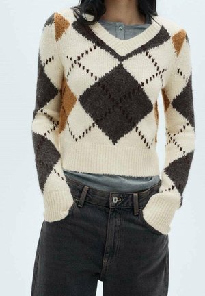 Vrouw draagt een crème-, bruin- en oranje geblokt V-hals trui over een grijs shirt, gecombineerd met hooggetailleerde zwarte jeans met haar handen in de zakken.