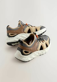 Des baskets d'extérieur en tissu beige, cuir marron, accents gris, ventilation sur les côtés et semelles blanches mouchetées avec des crampons noirs.