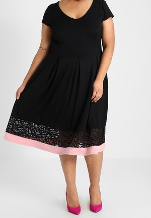 Kvinde iført en sort knælængde kjole med korte ærmer og en lyserød kant, kombineret med lyserøde højhælede sko, der holder i kjolens skørt.