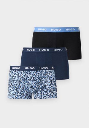 Atletyczny czarnoskóry mężczyzna stoi boso, zwrócony w prawo, nosi granatowe bokserki z paskiem z logo "Hugo", na jasnym, jednolitym tle.