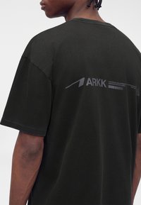 Άτομο που φοράει μαύρο t-shirt με την επωνυμία ARKK, εμφανιζόμενο από πίσω και πλάγια, με κοντά πλεγμένα μαλλιά σε μονόχρωμο φόντο.
