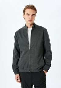 Grå zip-up jakke med høj krave, elastiske manchetter og talje. Lavet af et tekstureret stof; har en glat lynlås og rene linjer.