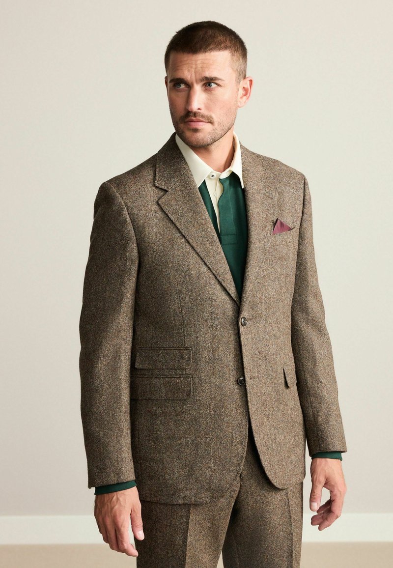 Brown tweed suit with notched lapels and two buttons. Features patch pockets, a green inner shirt, and a purple pocket square. Neat fit.
