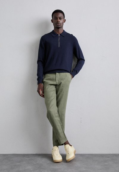 Marine blauwe polo met rits, lichtgroene broek en crèmekleurige sneakers. Model staat met ontspannen armen tegen een gestructureerde grijze achtergrond.