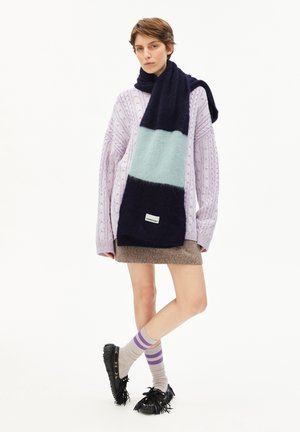 Jong persoon die een oversized lavendel kabelgebreid trui draagt, een kleurblok sjaal in marineblauw en lichtblauw, een bruine minirok, gestreepte sokken en zwarte franjesloafers.