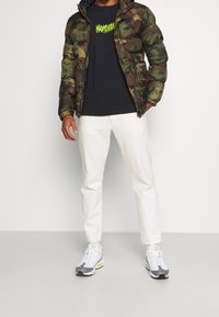 Camouflage puffer jacket, black graphic t-shirt, white trousers, and gray sneakers with neon accents. Visible zippers and textured fabric.