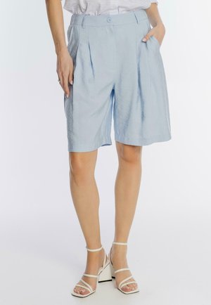 Kobieta w jasnoniebieskich plisowanych szortach do kolan i białych sandałach na słupku z paskami, stojąca z rękami częściowo w kieszeniach.