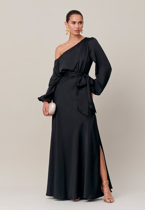 Femme en longue robe noire à une épaule avec manches longues, fente latérale, boucles d'oreilles dorées, tenant une petite pochette blanche et portant des sandales dorées.