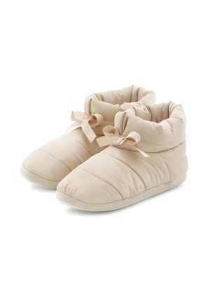 Beige padded slippers with a soft fabric upper, elasticized ankle, and decorative bows on each side. Textured sole provides grip.