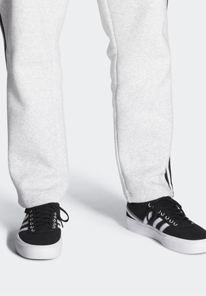 Jasnoszare dresy o miękkiej fakturze z czarnymi paskami po bokach, w zestawieniu z czarnymi sneakersami, które mają białe podeszwy i sznurówki.