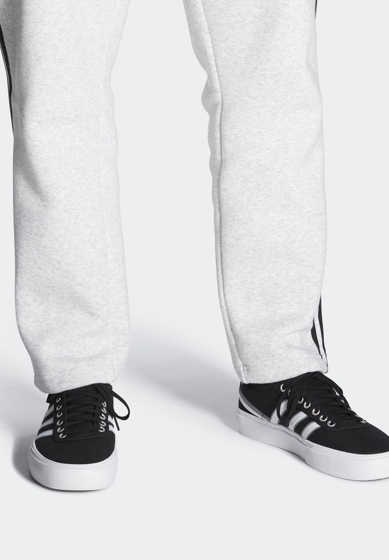 Harmaat collegehousut pehmeällä pinnalla ja mustilla raidoilla sivuilla, yhdistettynä mustiin lenkkikenkiin, joissa on valkoiset pohjat ja nauhat.