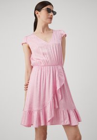 Pink dress with short sleeves, v-neck, gathered waist, and ruffled hem. Soft fabric with subtle pattern. Model wearing sunglasses.