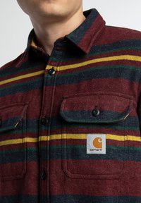 Camisa de franela a cuadros en rojo y azul con rayas verdes y amarillas, botones negros, dos bolsillos en el pecho y un parche con el logotipo de Carhartt.