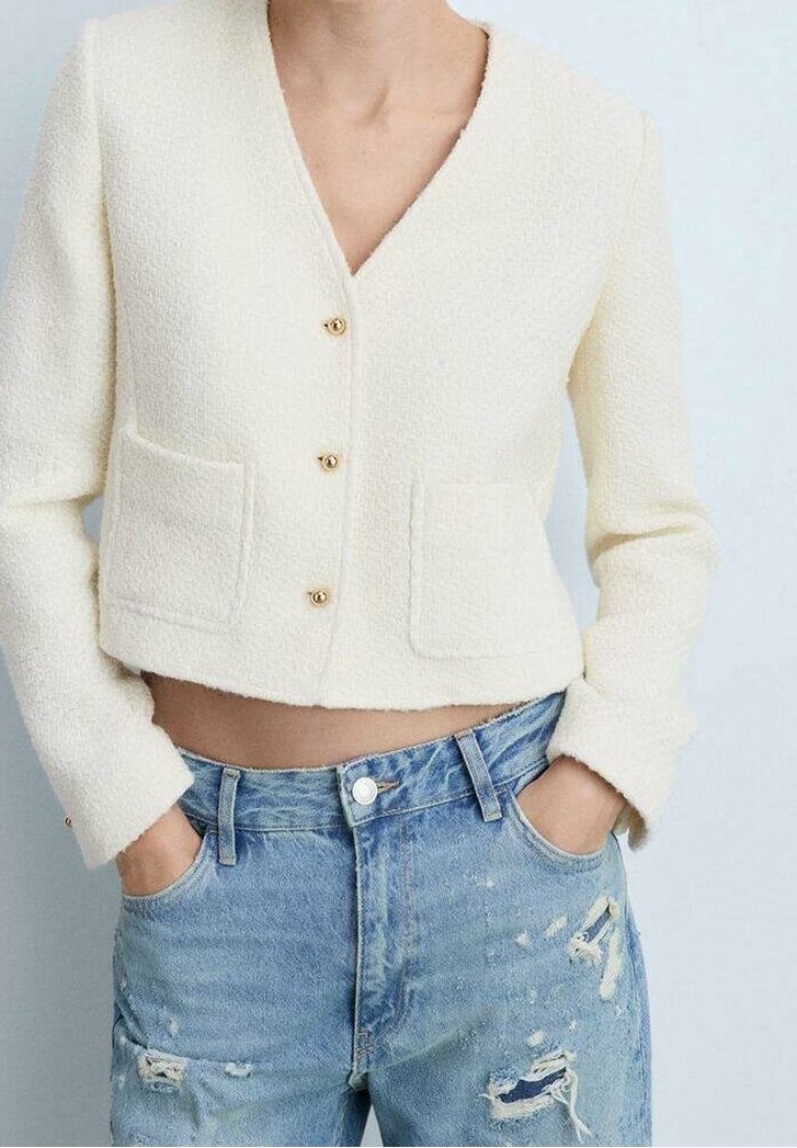 Cropped, cream textured jacket with a V-neck and two front pockets, featuring gold buttons. Paired with distressed light blue jeans.