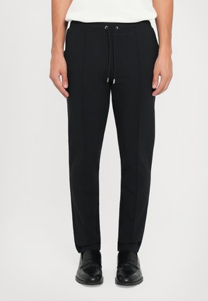 Black tailored pants made from smooth fabric with a drawstring waistband, front pleats, and slim fit, paired with black shoes.