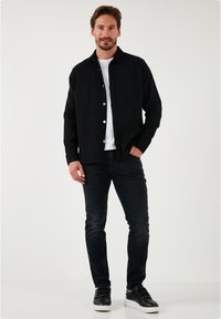 Chaqueta de mezclilla negra con botones blancos, bolsillo frontal, sobre una camiseta blanca, combinada con jeans negros y zapatillas negras.