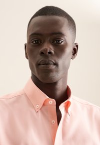 Camicia button-up rosa chiaro con un tessuto texture, caratterizzata da un colletto standard e dettagli con bottoni, indossata da un modello maschile su uno sfondo neutro.