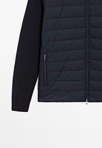 Puffer jacket with a navy quilted front and dark knit sleeves, featuring a full zipper and side zippered pockets. Smooth texture.