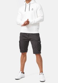 Homme portant un sweat à capuche blanc, un short cargo noir, des baskets blanches, debout les mains jointes devant sur fond blanc.