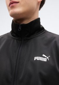 Μαύρο αθλητικό μπουφάν με ψηλό γιακά, μπροστινό φερμουάρ και λευκό λογότυπο Puma στο αριστερό στήθος. Το υλικό φαίνεται λείο και ελαφρύ.