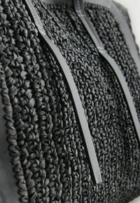 Sac tissé noir présentant une surface texturée, avec deux poignées en cuir noir lisse et des coutures visibles, soulignant son design artisanal.