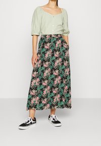 Femme portant un chemisier à manches bouffantes vert clair et une longue jupe noire à fleurs, avec des feuilles vertes et des fleurs roses, assortie de baskets noires.